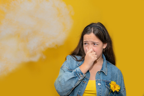 young woman in front of yellow wall holding her nose as a puff of smoke moves towards her